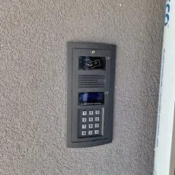 Szary panel domofonu z klawiaturą numeryczną i czytnikiem kart zbliżeniowych, zamontowany na szarej, chropowatej ścianie zewnętrznej budynku, obok pionowej rury osłonowej.