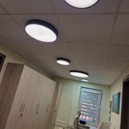 Równomiernie rozmieszczone, okrągłe lampy sufitowe LED w biurze z szafami w tle i oknem z żaluzjami.