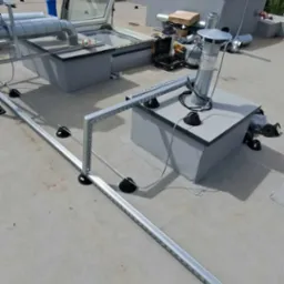 Widok z góry na płaski dach z zamontowanymi elementami wentylacji, kominkami, świetlikiem dachowym i aluminiową konstrukcją wsporczą, prawdopodobnie pod instalacje. Widoczne narzędzia i materiały...