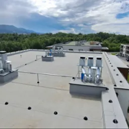 Instalacja odgromowa na płaskim dachu nowego budynku mieszkalnego z widocznymi uchwytami, przewodami i zwodami pionowymi, na tle zieleni i gór.