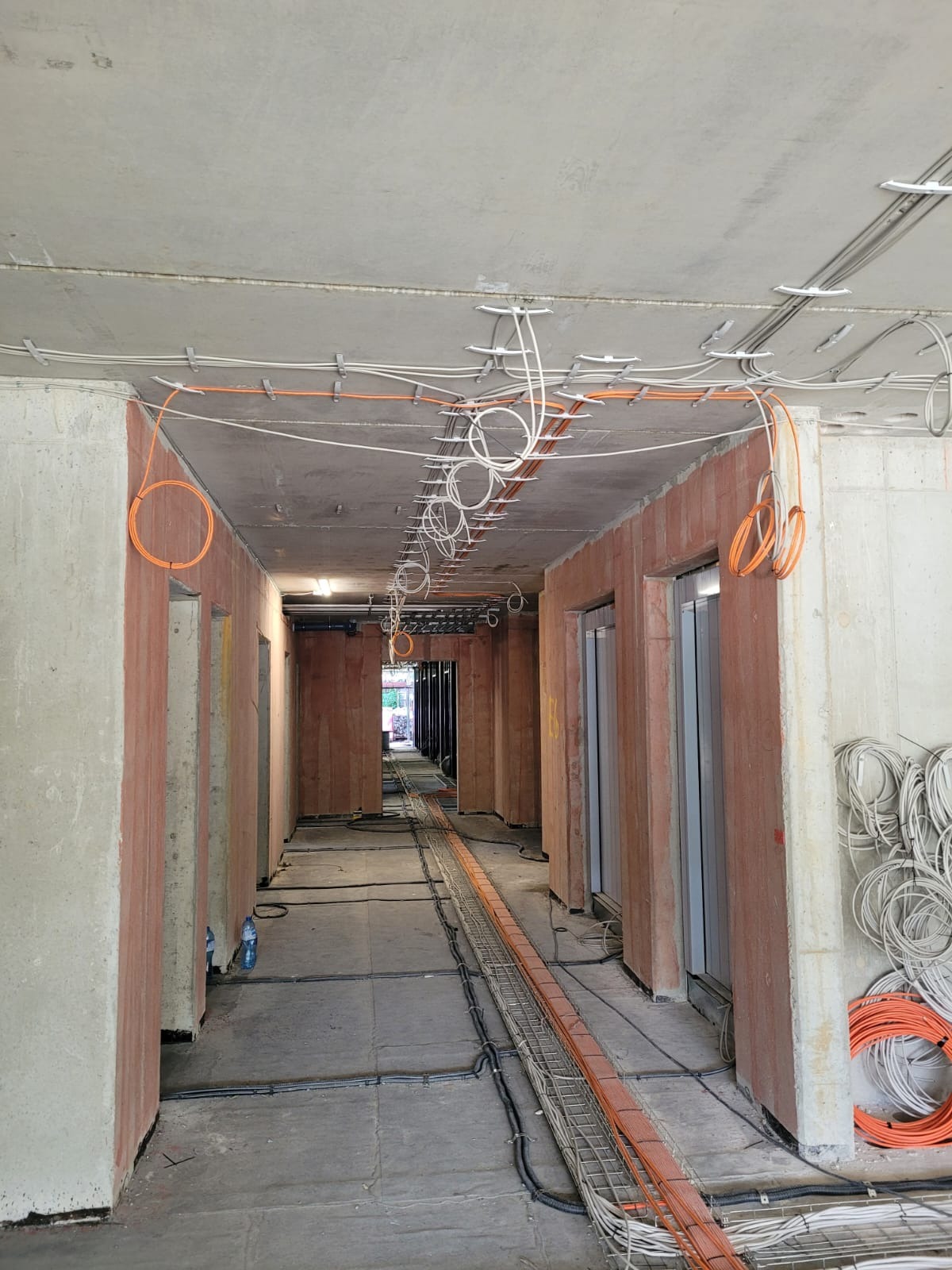 Korytarz w stanie surowym z widocznymi przewodami elektrycznymi prowadzonymi pod sufitem i w korytach kablowych na podłodze, przewody w kolorze białym i pomarańczowym, ściany w surowym betonie.