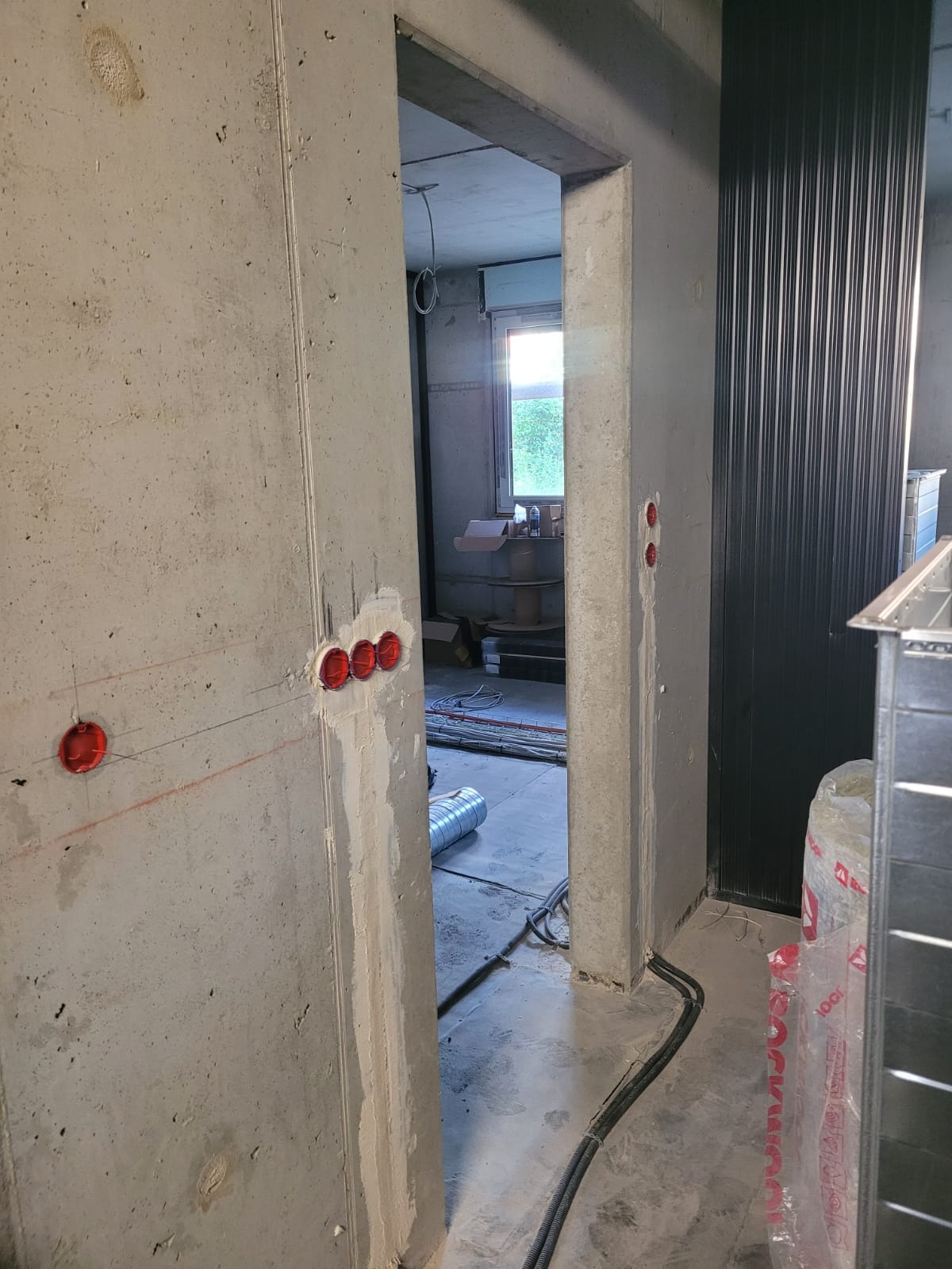 Surowy betonowy mur z trzema czerwonymi puszkami instalacyjnymi, widoczne przewody i elementy wentylacyjne w tle, wnętrze w trakcie prac wykończeniowych.