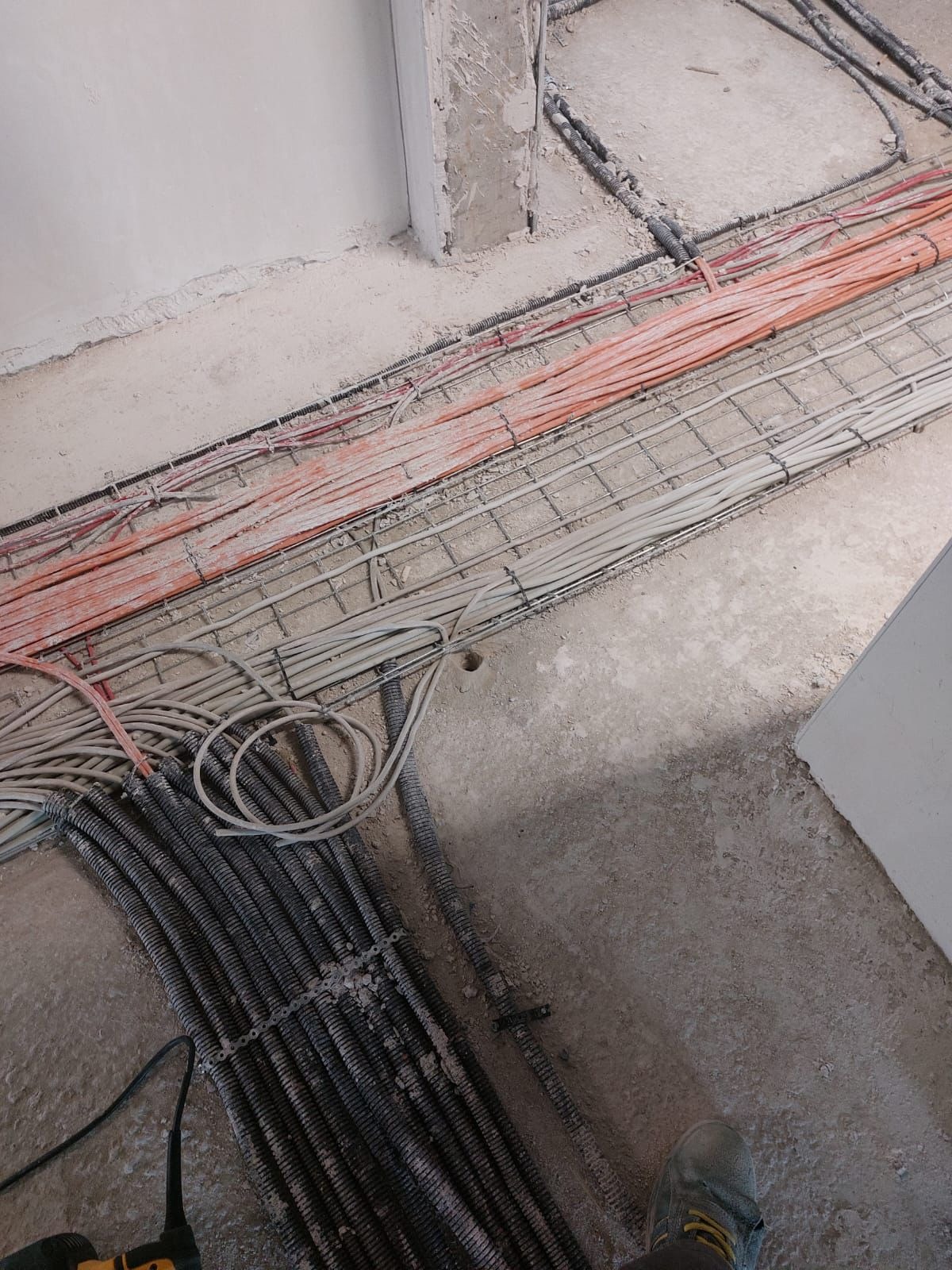 Układanie przewodów elektrycznych w korytach kablowych na surowym podłożu, widoczne peszle i kable różnej grubości, w tle fragment ściany w trakcie prac wykończeniowych.