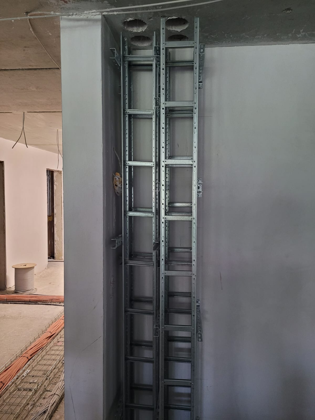 Metalowa konstrukcja koryt kablowych zamontowana pionowo na ścianie w surowym stanie, widoczne otwory w suficie na przewody, w tle bęben z kablem i ułożone kable w korycie.