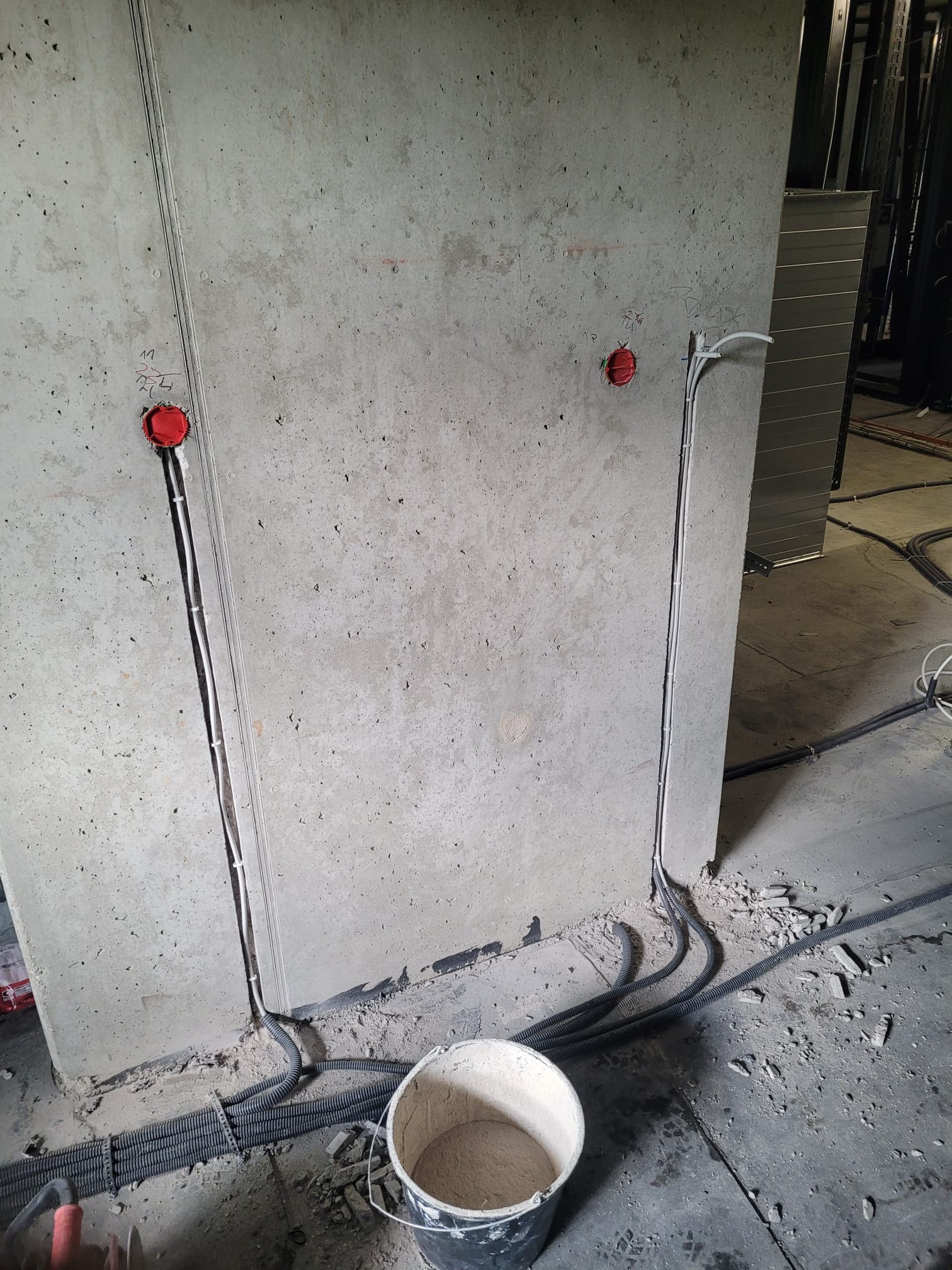 Betonowa ściana z wyżłobionymi kanałami, w których umieszczono przewody elektryczne zakończone czerwonymi puszkami. Na podłodze widoczne gruz i rury karbowane oraz wiadro z zaprawą.