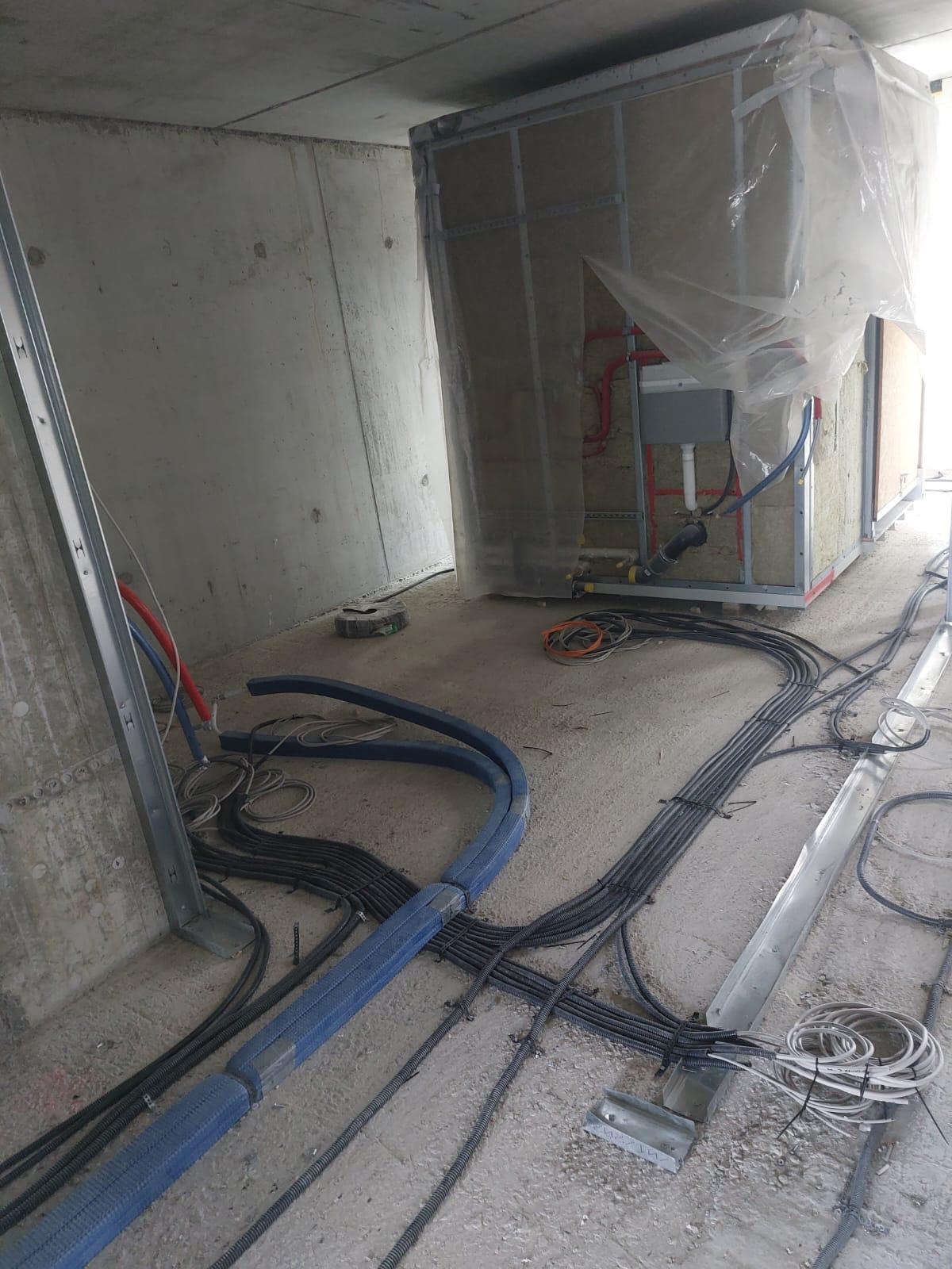 Surowy stan instalacji elektrycznych w budynku w trakcie budowy, widoczne kable w peszlach i rurach, skrzynka instalacyjna w osłonie z wełny mineralnej i folii, betonowe ściany i podłoga.