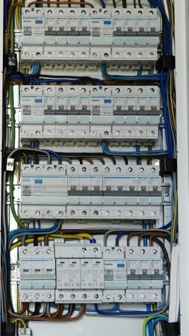 Rozdzielnia elektryczna z zamontowanymi bezpiecznikami różnicowoprądowymi i przewodami w kolorach niebieskim, żółto-zielonym i brązowym, widoczne oznaczenia producenta Hager.