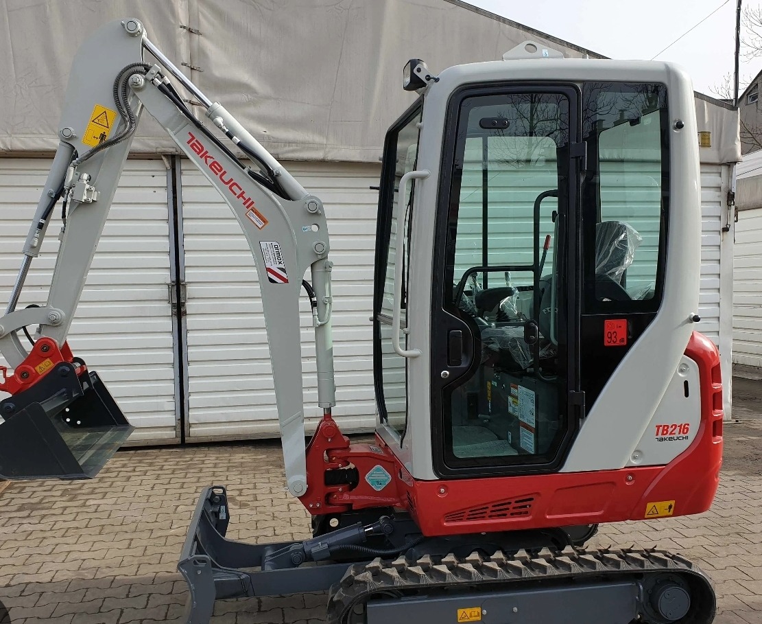 Nowa, szaro-czerwona mini koparka Takeuchi TB216 z kabiną, zaparkowana na kostce brukowej przed białym magazynem.