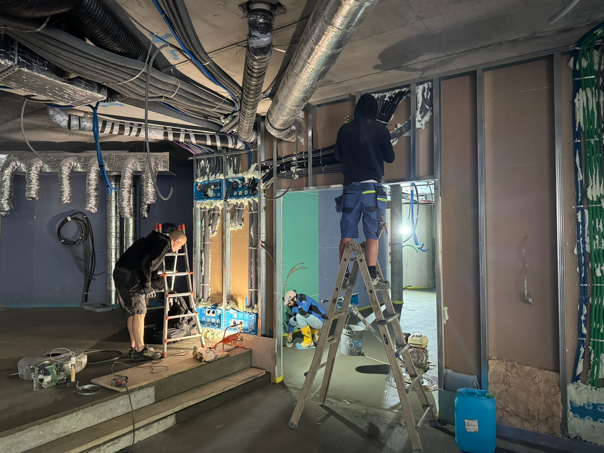 Montaż instalacji w surowym wnętrzu: Dwóch elektryków na drabinach układa kable w ścianach z profili, widoczne liczne przewody pod sufitem i na ścianach, narzędzia i materiały budowlane wokół.