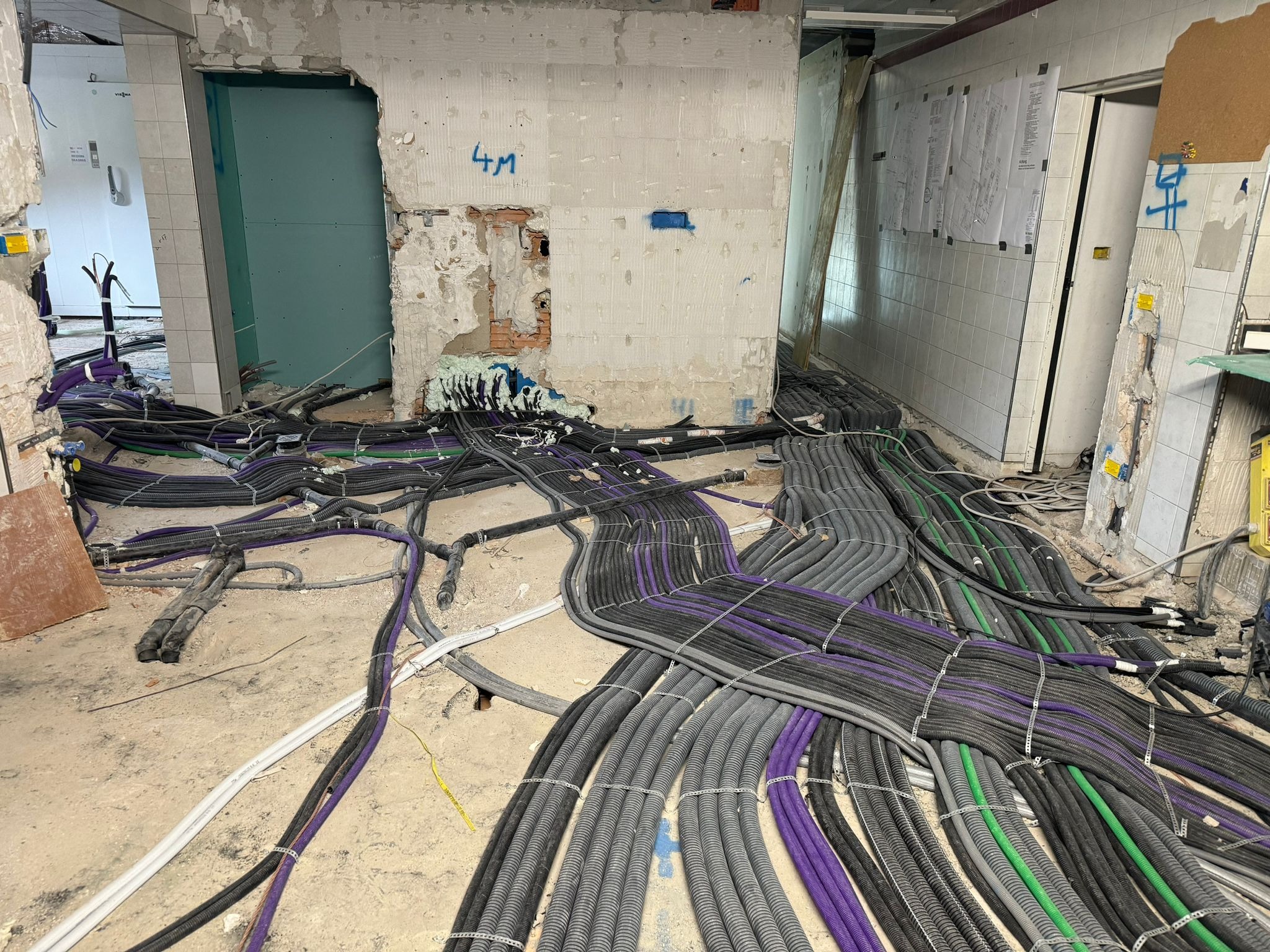 Rzędy przewodów elektrycznych w peszlach, częściowo fioletowe, szare i zielone, ułożone na surowej podłodze w trakcie remontu, ściany w stanie deweloperskim z widocznymi instalacjami i planami...