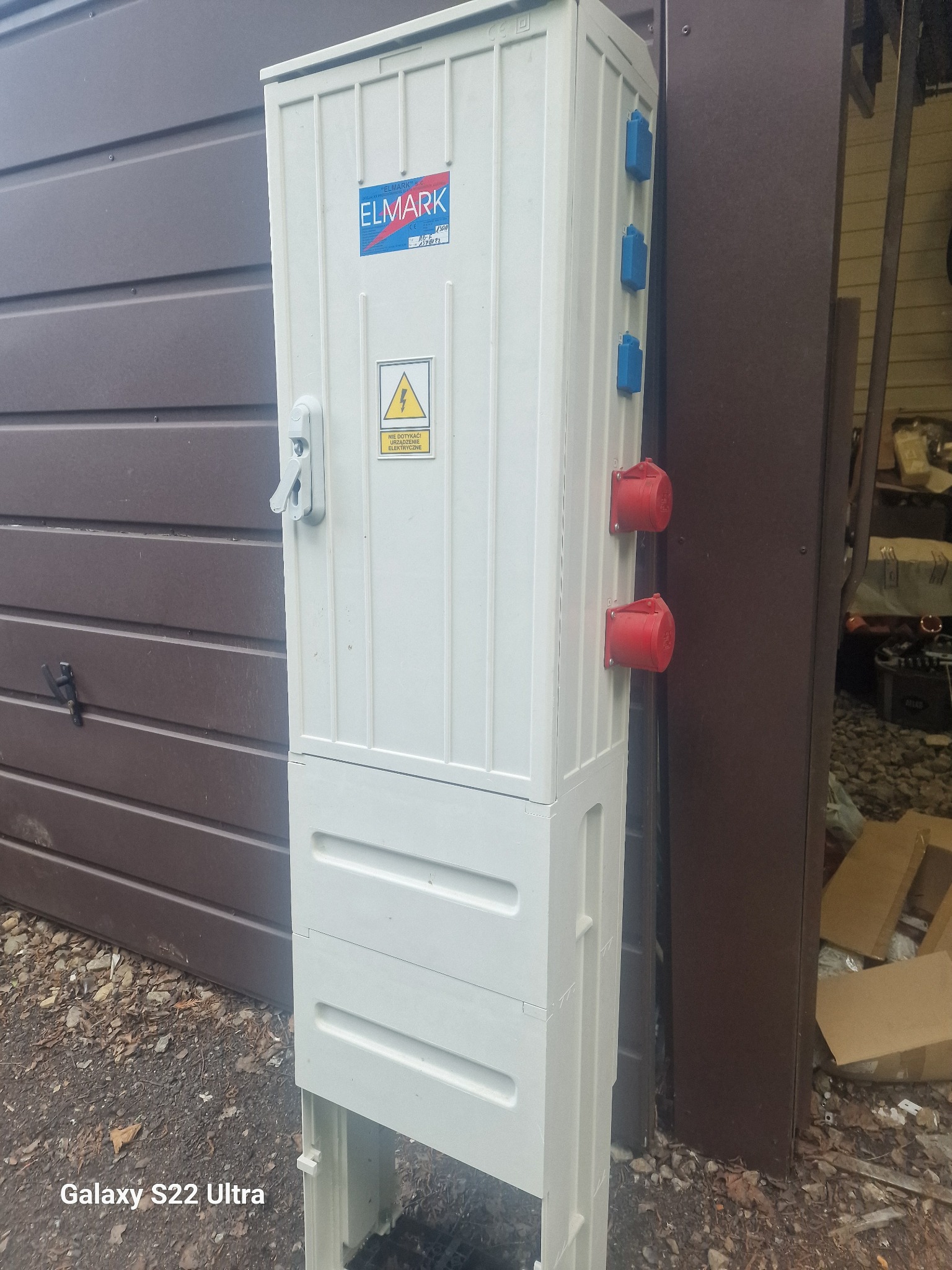 Biała szafa elektryczna z gniazdami siłowymi (czerwone) i standardowymi (niebieskie), umieszczona na zewnątrz budynku, z widocznym znakiem ostrzegawczym o napięciu elektrycznym i logo firmy ELMARK.
