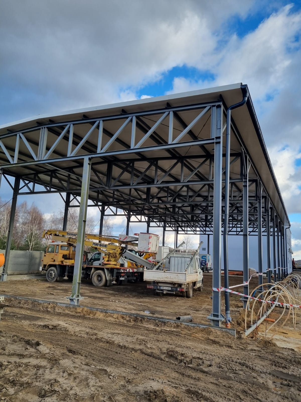 Konstrukcja stalowa zadaszenia w trakcie montażu, widoczne dźwigi i samochody dostawcze na placu budowy, z instalacją odgromową na dachu, w tle drzewa i pochmurne niebo.