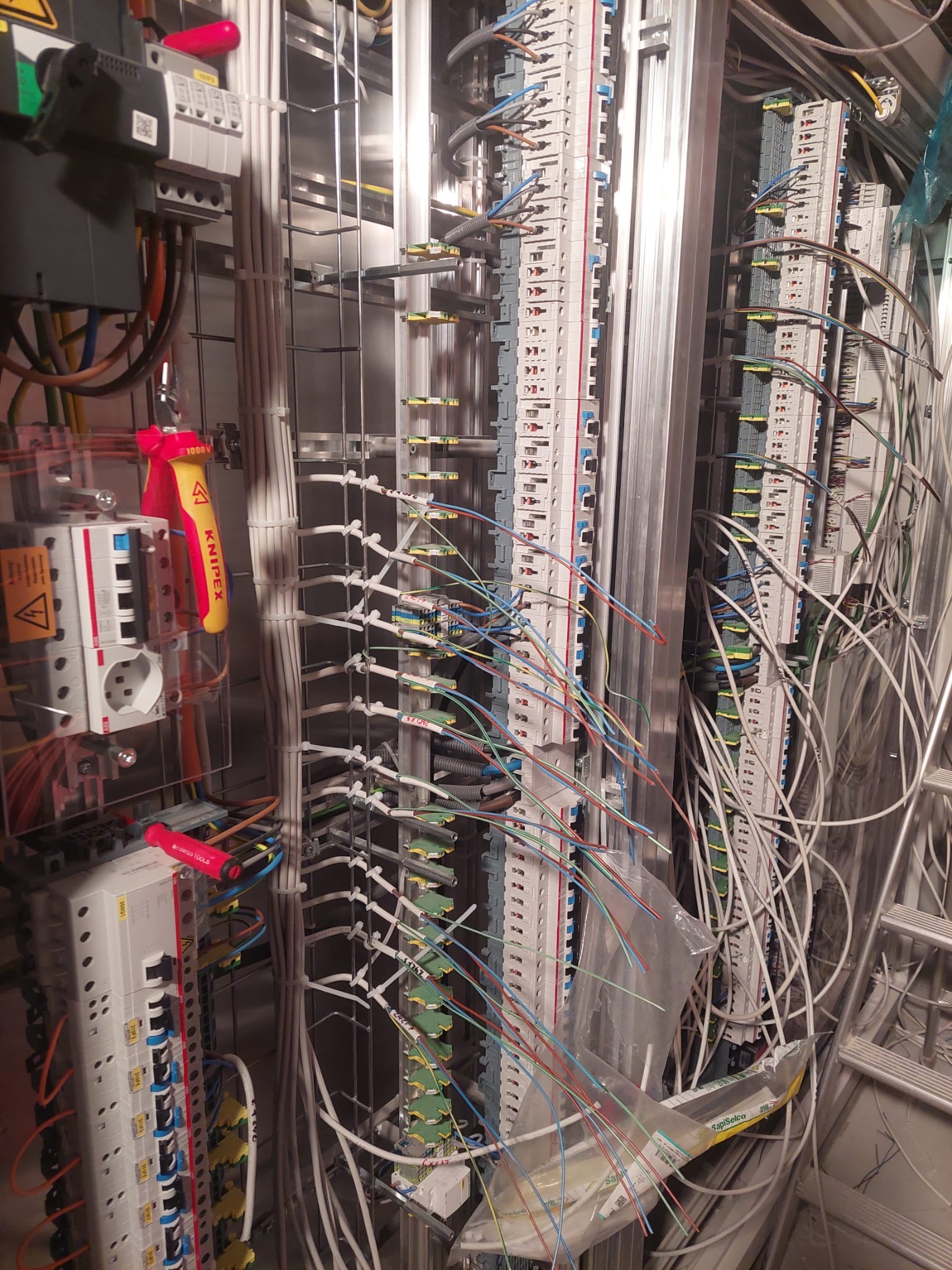 Rozdzielnica elektryczna w trakcie montażu, widoczne liczne przewody, listwy zaciskowe, zabezpieczenia, narzędzia Knipex i Swiss Tools, oraz drabina.