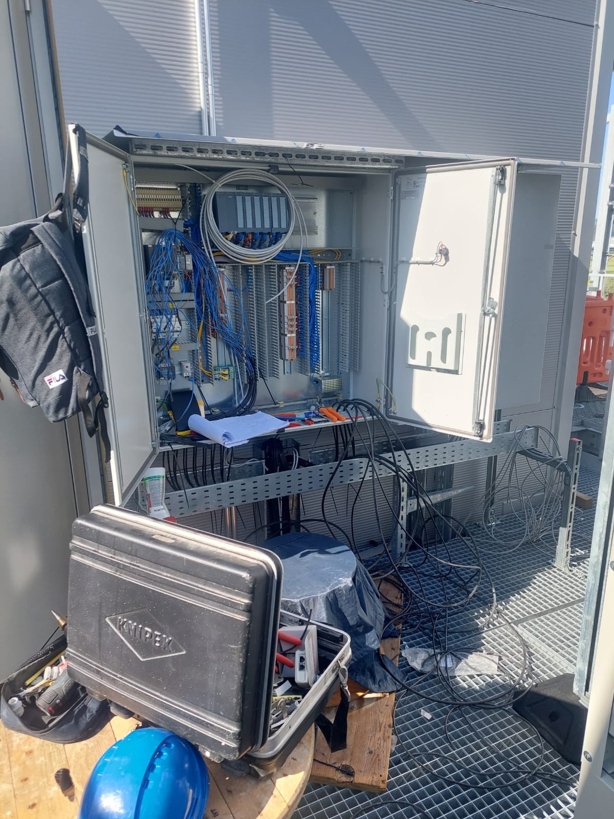 Otwarta szafa sterownicza z widocznymi przewodami, listwami zaciskowymi i modułami sterującymi, narzędzia i dokumentacja robocza na zewnątrz szafy, plecak Fila zawieszony na boku szafy.