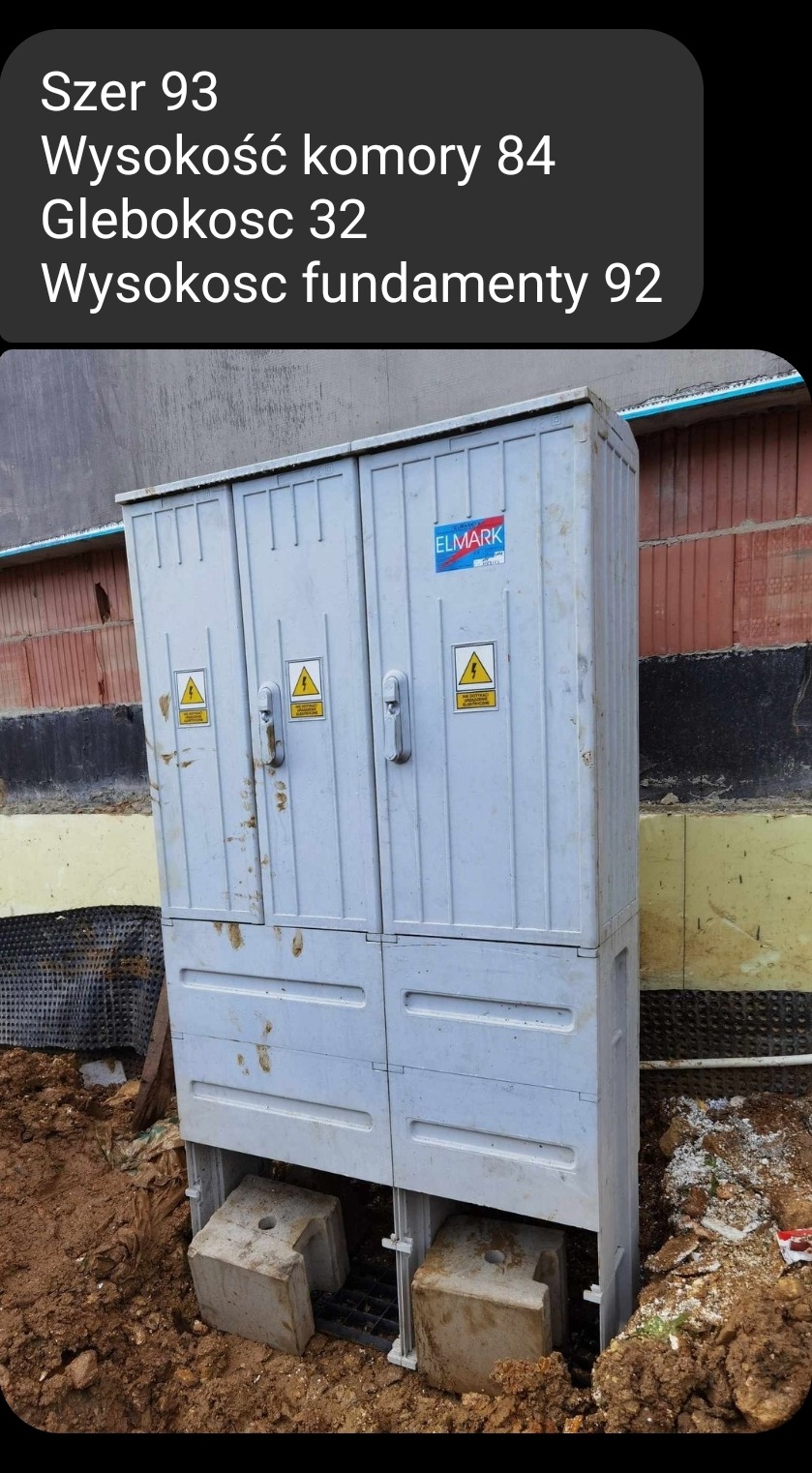 Szara, zewnętrzna szafa elektryczna z naklejkami ostrzegawczymi, umieszczona na betonowych fundamentach w wykopie ziemnym obok ściany budynku w trakcie budowy.