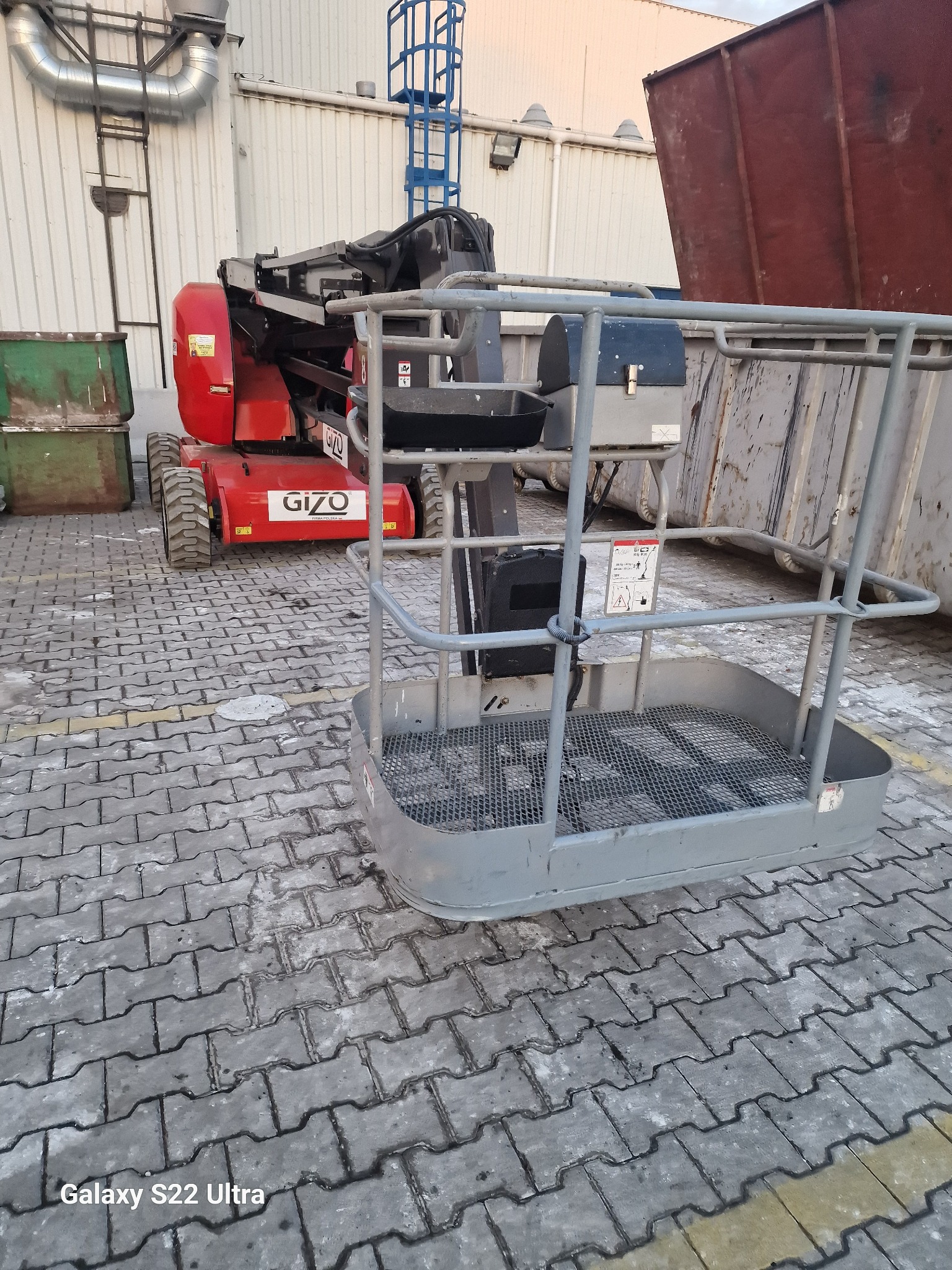 Czerwony podnośnik koszowy Gizo na kostce brukowej przed halą przemysłową i kontenerami.