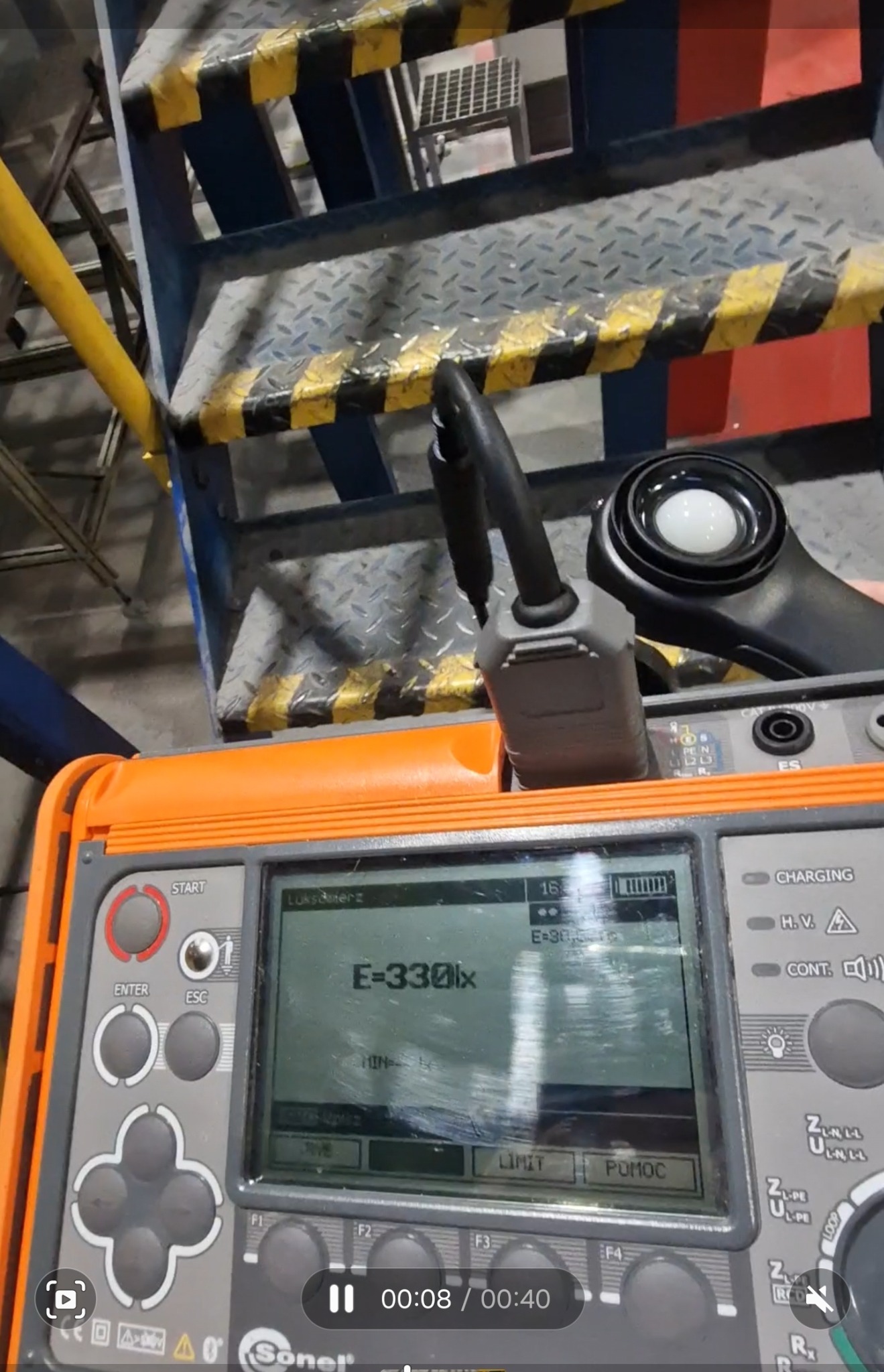 Miernik natężenia światła Sonel wskazuje wartość 330 lx, umieszczony na tle metalowych schodów z żółto-czarnymi pasami ostrzegawczymi.