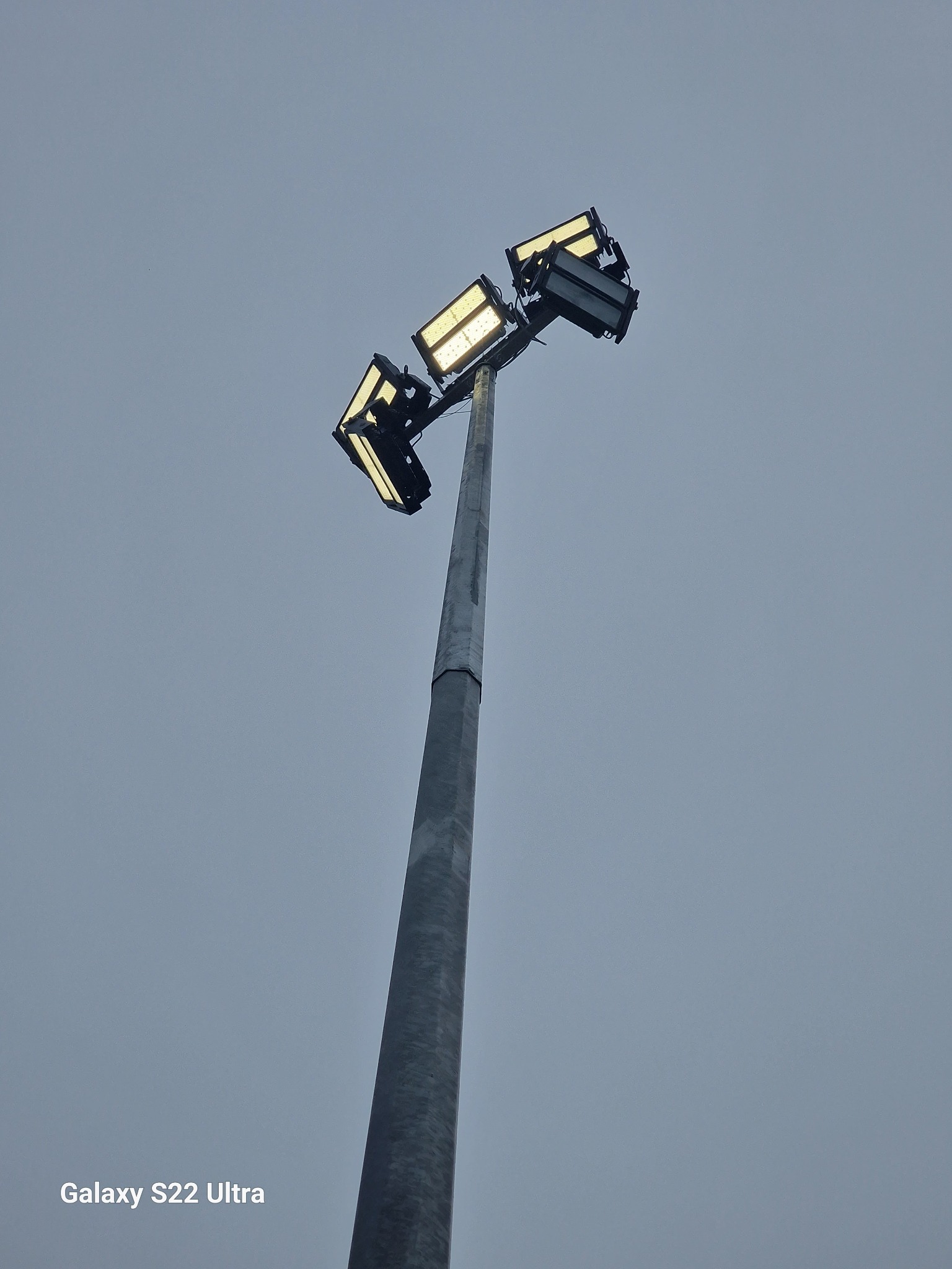 Wysoki maszt oświetleniowy z czterema reflektorami LED skierowanymi w różnych kierunkach, sfotografowany pod kątem w pochmurny dzień.