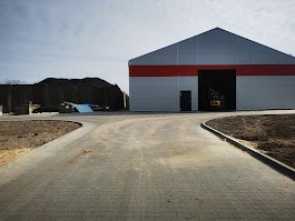 Szara, betonowa nawierzchnia z kostki brukowej prowadząca do nowoczesnego magazynu z czerwoną linią. Po lewej pryzma materiału budowlanego.