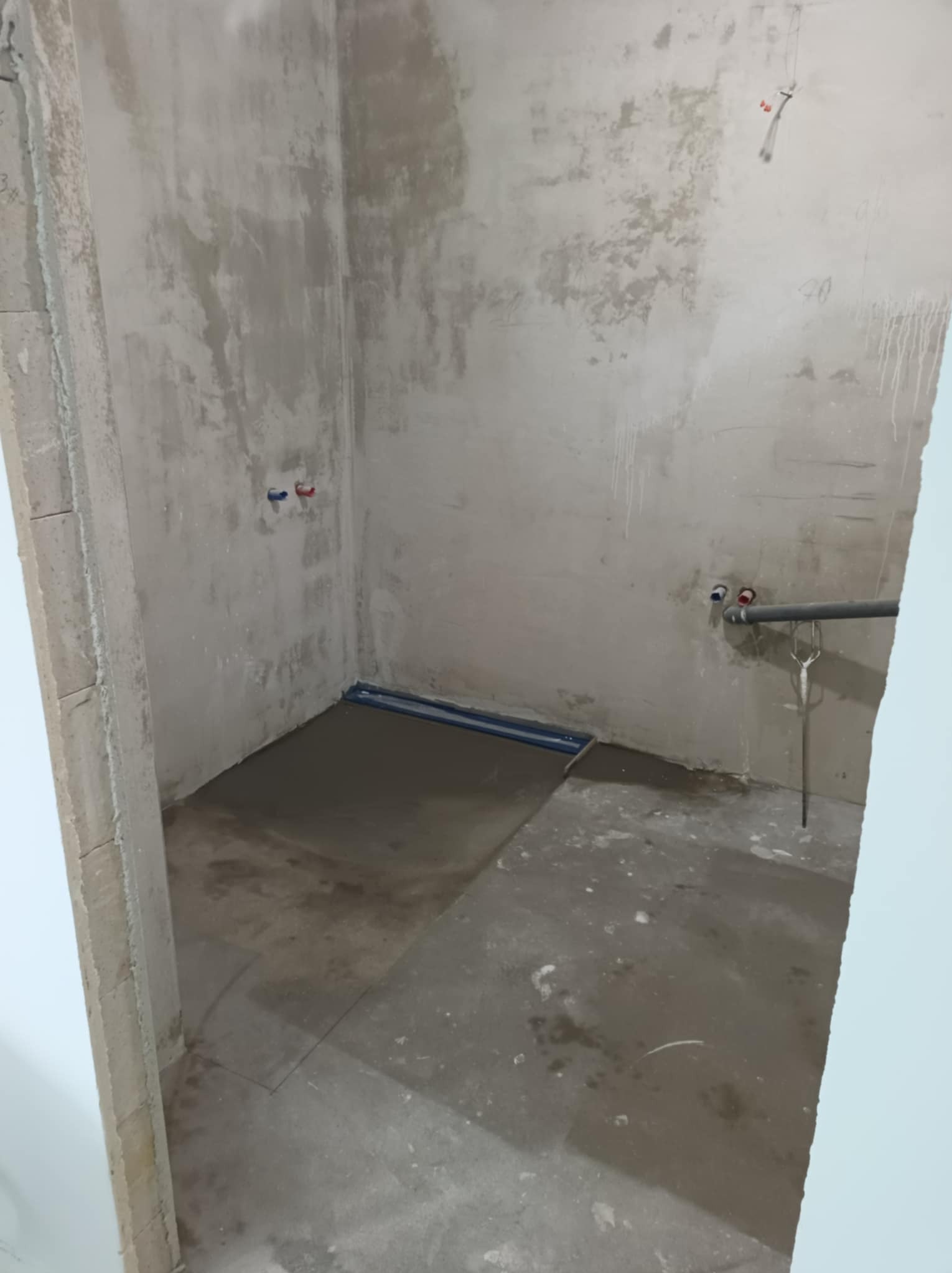 Surowy stan łazienki w trakcie remontu: wylewka podłogowa, odpływ liniowy, surowe ściany z widocznymi instalacjami wodnymi. Wnętrze w trakcie prac wykończeniowych.