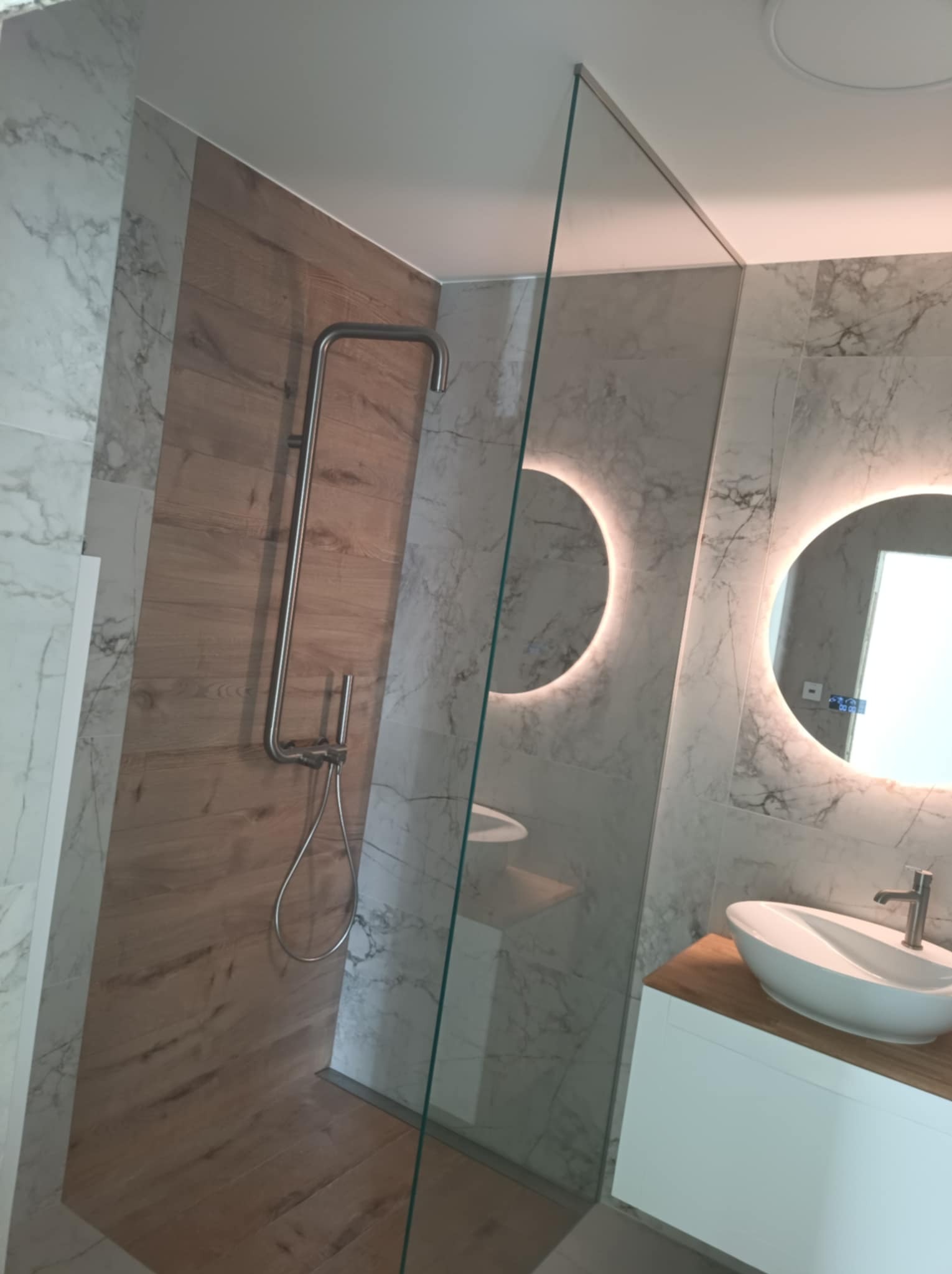 Nowoczesna łazienka z kabiną prysznicową typu walk-in, drewnopodobnymi płytkami i okrągłym lustrem z podświetleniem LED. Minimalistyczny design, jasne marmurowe płytki.