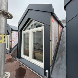 GB Dachy - Nowoczesne okna dachowe w grafitowej obudowie, zamontowane na dachu krytym blachą, widoczne z rusztowania.