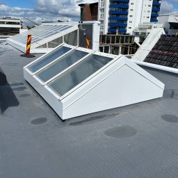 GB Dachy - Płaski dach pokryty szarą membraną z trzema białymi świetlikami, widoczne budynki w tle i elementy zabezpieczeń.