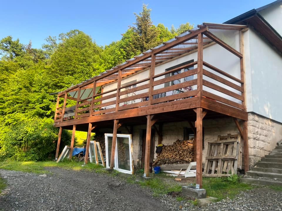 Do budynku dobudowany drewniany taras z zadaszeniem z przezroczystego tworzywa, widoczne przygotowane drewno i stare okna.