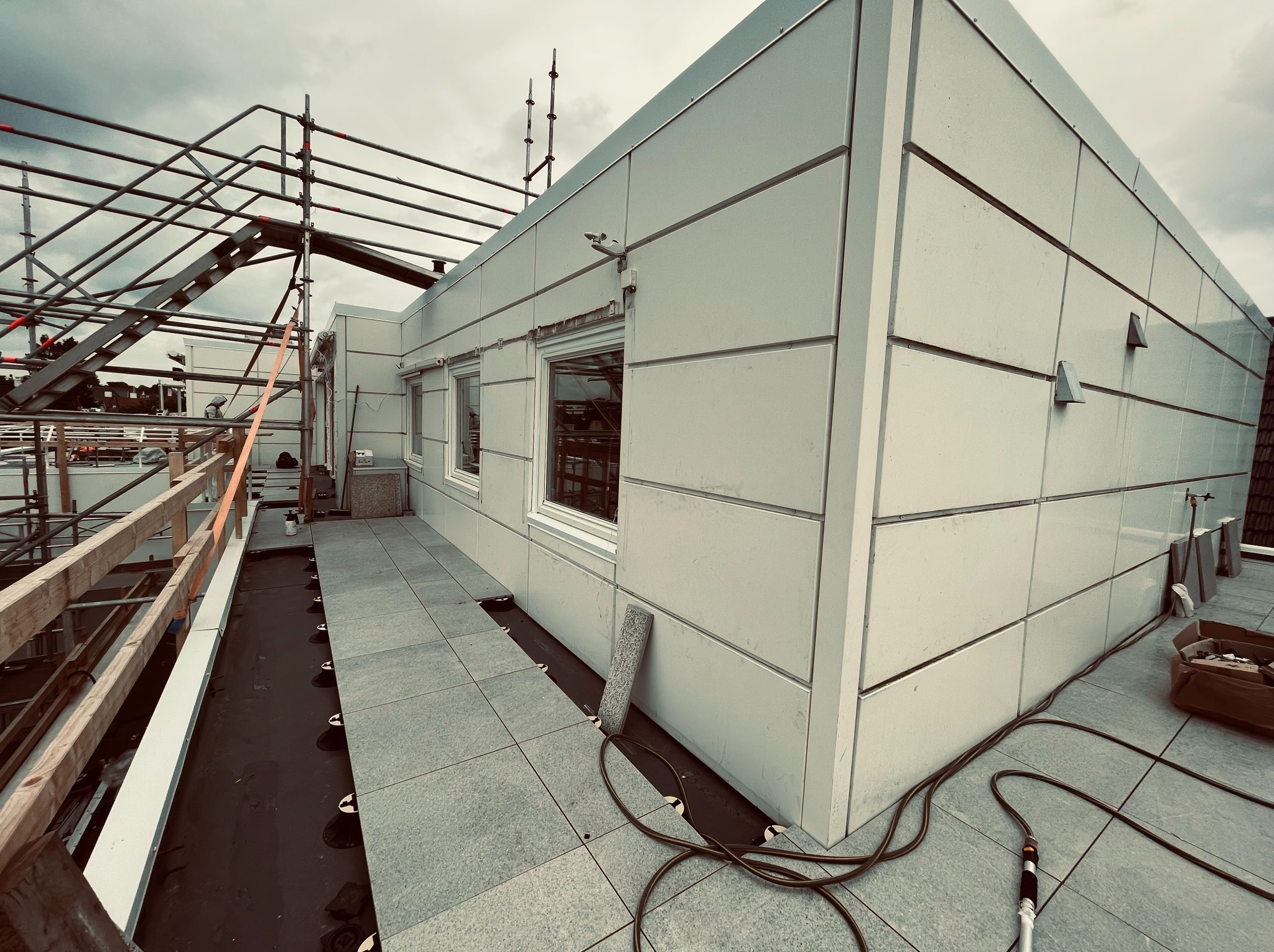 Nowoczesny dach płaski z białymi panelami elewacyjnymi i szarą kostką brukową, widoczny fragment rusztowania i przewody elektryczne na dachu.