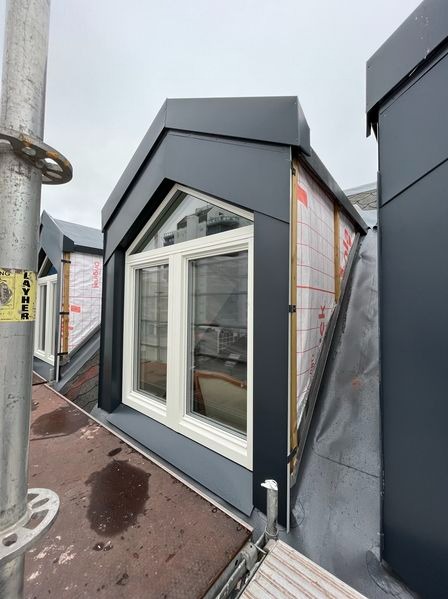 Nowoczesne okna dachowe w grafitowej obudowie, zamontowane na dachu krytym blachą, widoczne z rusztowania.