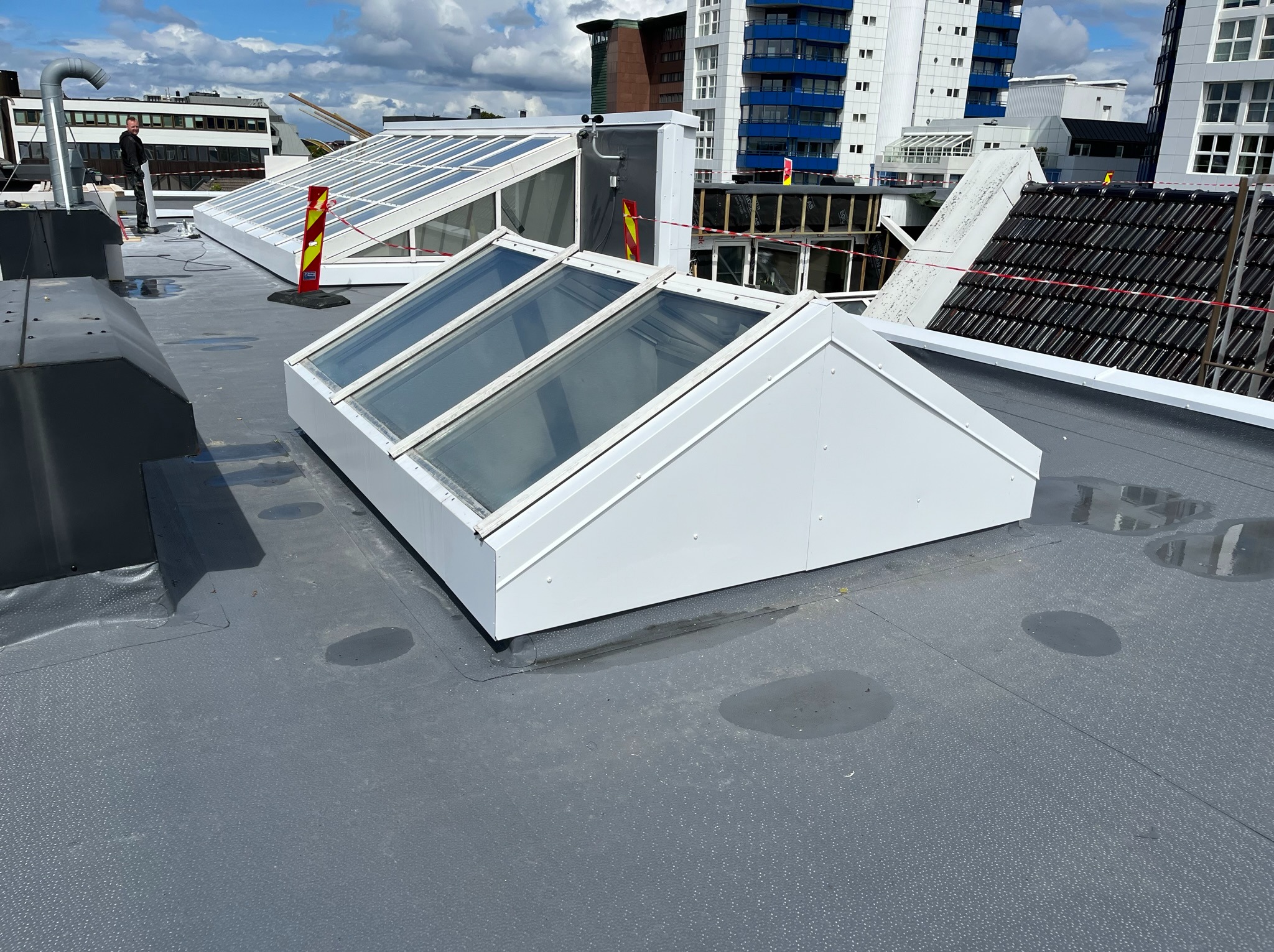 Płaski dach pokryty szarą membraną z trzema białymi świetlikami, widoczne budynki w tle i elementy zabezpieczeń.