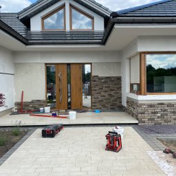 Wykończenia Wnętrz - Montaż okładziny z cegły klinkierowej na elewacji domu, widoczne narzędzia, poziomica laserowa i zapas materiału na palecie.