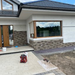 Wykończenia Wnętrz - Montaż elewacji z cegły klinkierowej na fragmencie domu jednorodzinnego, widoczne narzędzia i materiały budowlane, poziomica laserowa, fragment nowej kostki brukowej i przygotowanego terenu zielonego.