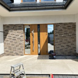 Wykończenia Wnętrz - Wykończenie elewacji wejściowej domu z użyciem cegły klinkierowej, widoczne drzwi wejściowe z elementami szkła oraz rozstawiona drabina aluminiowa.