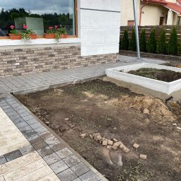 Wykończenia Wnętrz - Układanie kostki brukowej na posesji, widoczne fragmenty ułożonego chodnika z szarej i beżowej kostki, przygotowane rabaty kwiatowe i fragment elewacji budynku z kwiatami w doniczkach.