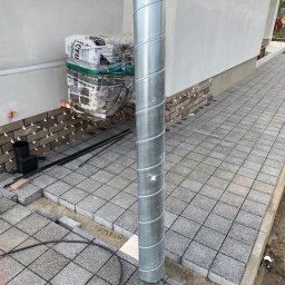 Wykończenia Wnętrz - Montaż elewacji z cegły klinkierowej i układanie szarej kostki brukowej na posesji przy budynku, widoczne paczki z klejem i dystanse krzyżowe.