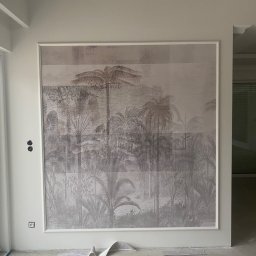 Wykończenia Wnętrz - Nowoczesny salon w trakcie wykańczania, z dużym panelem ściennym przedstawiającym tropikalny krajobraz w odcieniach szarości, widoczne gniazdka elektryczne i fragment podłogi z resztkami tapety.