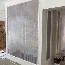 Wykończenia Wnętrz - Ściana z dekoracyjną, szaro-złotą tapetą w pionowe pasy, imitującą krajobraz górski, widoczna w trakcie remontu, z kartonem po lodówce Samsung w tle i narzędziami malarskimi w pobliżu