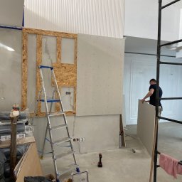 Wykończenia Wnętrz - Montaż płyty gipsowo-kartonowej na drewnianej konstrukcji w trakcie remontu, widoczna drabina, narzędzia i pracownik w tle.