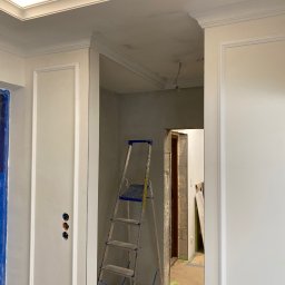 Wykończenia Wnętrz - Wnętrze w trakcie prac wykończeniowych: białe ściany z dekoracyjnymi listwami, aluminiowa drabina w przejściu, zabezpieczone folią drzwi.