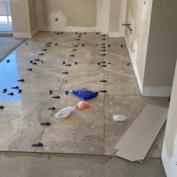 Wykończenia Wnętrz - Układanie jasnych, marmurkowych płytek podłogowych z użyciem systemu poziomowania; widoczny pracownik w roboczym stroju, niebieska torba z elementami systemu,  surowe ściany w tle.