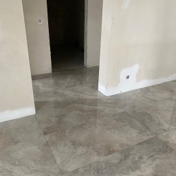 Wykończenia Wnętrz - Wnętrze w trakcie prac wykończeniowych: świeżo położona, błyszcząca, szara terakota imitująca marmur, ściany po gruntowaniu z widocznymi miejscami po szpachlowaniu gipsem wokół gniazdek...