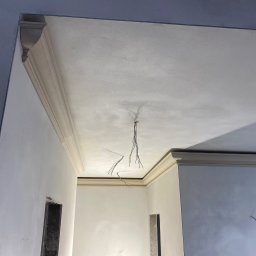 Wykończenia Wnętrz - Świeżo pomalowany sufit z dekoracyjną listwą przysufitową w jasnym odcieniu, widoczne wystające przewody elektryczne, ściany przygotowane do malowania w odcieniach szarości i bieli, widoczne...
