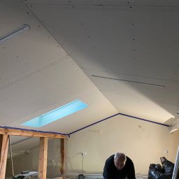 Wykończenia Wnętrz - Montaż płyt kartonowo-gipsowych na poddaszu z oknem dachowym; widoczne profile, wkręty i częściowo ocieplona ściana.