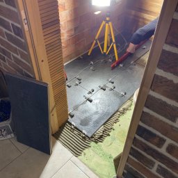 Wykończenia Wnętrz - Układanie ciemnych płytek podłogowych w pomieszczeniu z ceglanymi ścianami, widoczne krzyżyki dystansowe, poziomica i lampa na statywie.