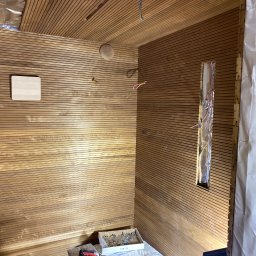 Wykończenia Wnętrz - Wnętrze pomieszczenia w trakcie wykończenia drewnem: poziome panele na ścianach i suficie, widoczne przewody elektryczne i puszka instalacyjna, narzędzia i elementy montażowe w kartonie na podłodze.