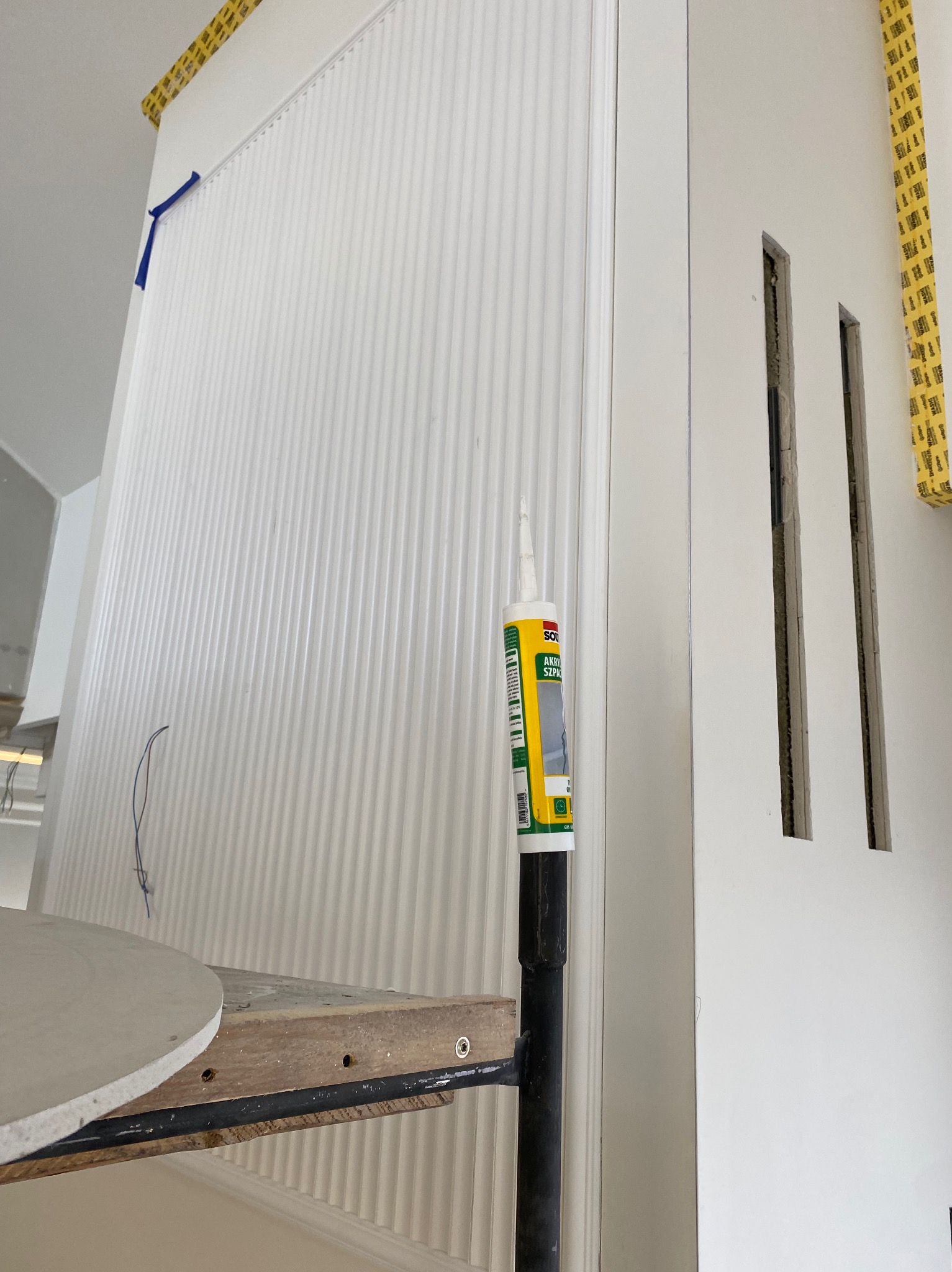Montaż dekoracyjnych paneli ściennych z pionowymi żłobieniami, widoczna tuba akrylu szpachlowego, fragment rusztowania i otwory w ścianie przygotowane pod instalacje.