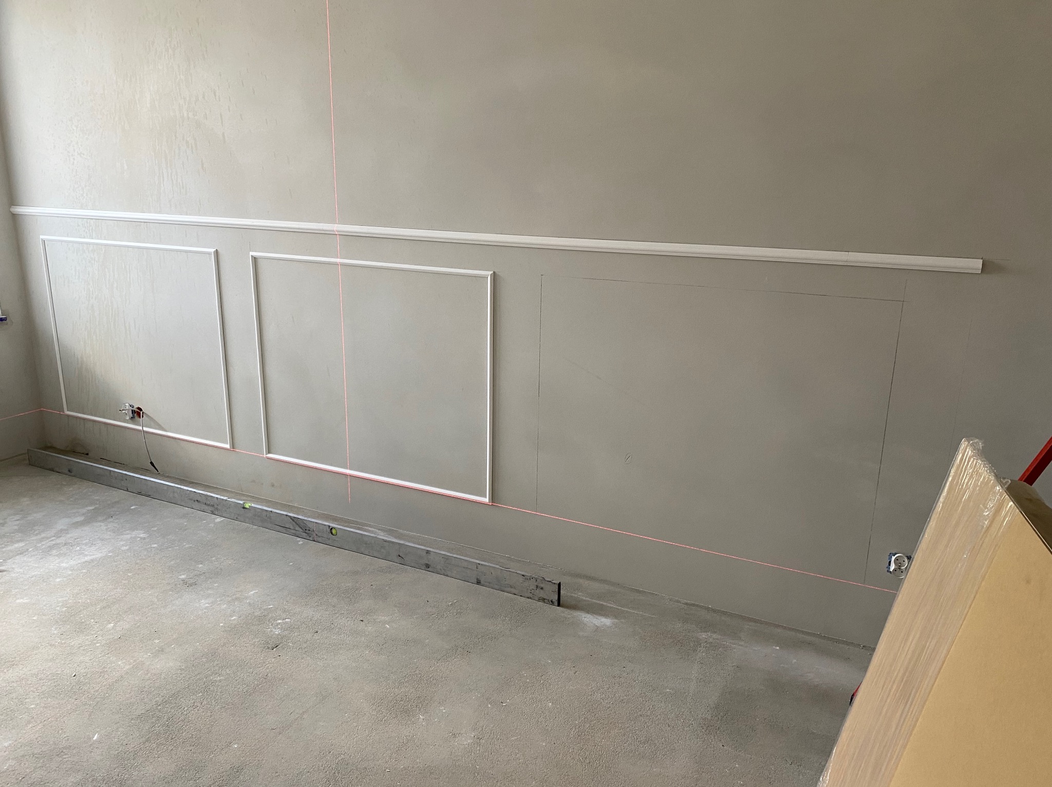 Ściana w trakcie dekorowania sztukaterią: zamontowane listwy przypodłogowe i ramy, wyznaczony laserem poziom, surowy beton na podłodze.