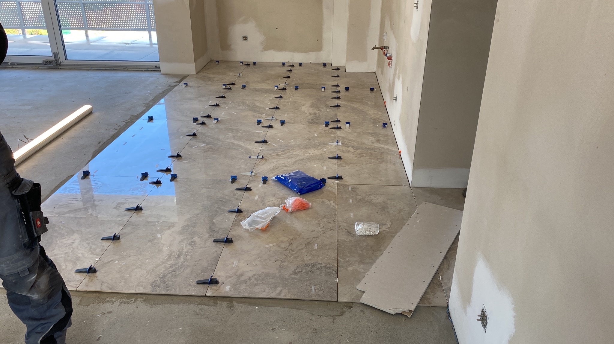 Układanie jasnych, marmurkowych płytek podłogowych z użyciem systemu poziomowania; widoczny pracownik w roboczym stroju, niebieska torba z elementami systemu,  surowe ściany w tle.