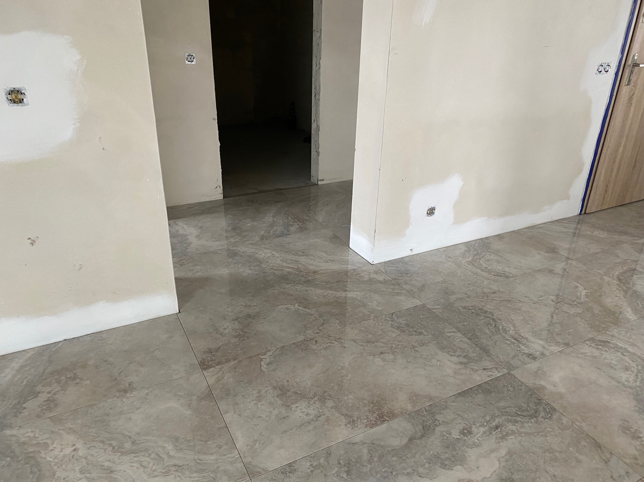 Wnętrze w trakcie prac wykończeniowych: świeżo położona, błyszcząca, szara terakota imitująca marmur, ściany po gruntowaniu z widocznymi miejscami po szpachlowaniu gipsem wokół gniazdek...